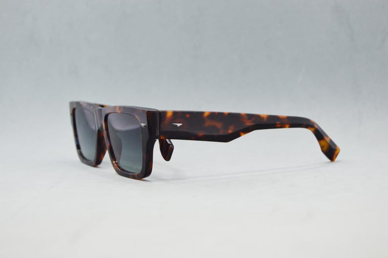 Puede incluir: Gafas de sol de carey con una montura gruesa y rectangular con patrones moteados en marr&oacute;n oscuro y &aacute;mbar. Las lentes son de color gris oscuro y las patillas son anchas, con un peque&ntilde;o detalle met&aacute;lico plateado cerca de la bisagra.