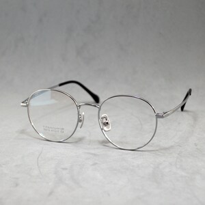 Könnte beinhalten: Silberfarbene runde Brille mit klaren Gläsern. Der Rahmen trägt den Text "TITANIUM 18  18K5 49□17 135 C2", der auf der Innenseite des linken Glases aufgedruckt ist.