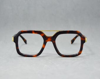 Puede incluir: Gafas de montura cuadrada con estampado de carey. Las gafas tienen un puente y detalles dorados en las patillas. Las lentes son transparentes y las monturas son una mezcla de marrón y negro.