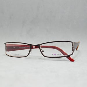 Könnte beinhalten: Braune Metallbrille mit roten Akzenten und klaren Gläsern. Der Rahmen trägt den Text "Stainless Steel" auf der linken Seite und "S-Vision" auf der rechten Seite.