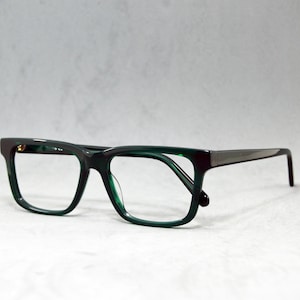 Könnte beinhalten: Dunkelgrüne Brille mit rechteckigem Rahmen und kontrastierenden dunkelgrauen Bügeln. Die Brille hat ein modernes Design und ist vor neutralem Hintergrund platziert. Die Gläser sind klar.