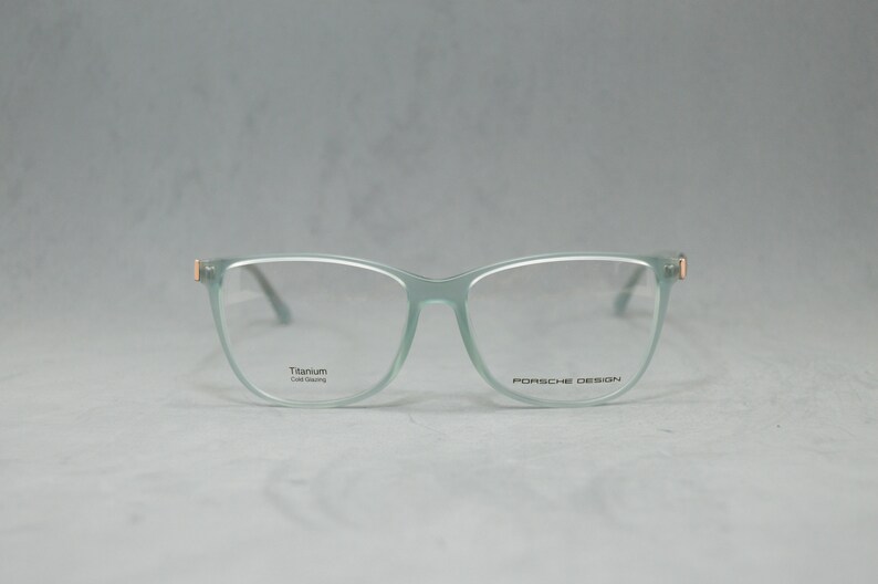 May include: A pair of light green eyeglasses with a titanium frame and gold accents. The glasses have a square shape with rounded edges and say "Porsche Design" on the right temple.