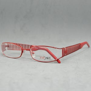 Può includere: Occhiali da vista con montatura in metallo rosso e lenti trasparenti. La montatura ha un design a righe e il marchio "Eye Art" è stampato sulla lente destra.