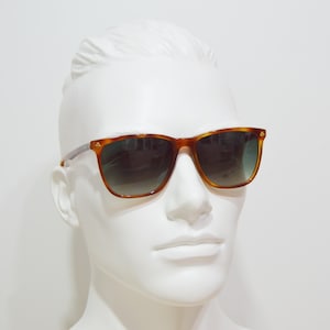 May include: A pair of brown tortoise shell sunglasses with green lenses. The sunglasses are on a white mannequin head.