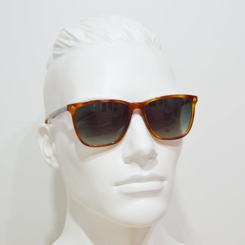 May include: A pair of brown tortoise shell sunglasses with green lenses. The sunglasses are on a white mannequin head.