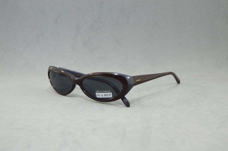 May include: Brown oval-shaped sunglasses with dark lenses. The sunglasses have a label that says "100% UV Protection" and "IN & OUT".