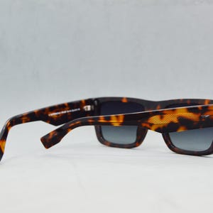 Puede incluir: Gafas de sol de carey con un patr&oacute;n moteado en marr&oacute;n oscuro y &aacute;mbar en las monturas y patillas gruesas. Las lentes rectangulares son de color gris oscuro con un tinte degradado. Un detalle texturizado es visible en la patilla exterior.