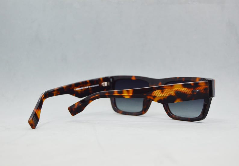 Puede incluir: Gafas de sol de carey con un patr&oacute;n moteado en marr&oacute;n oscuro y &aacute;mbar en las monturas y patillas gruesas. Las lentes rectangulares son de color gris oscuro con un tinte degradado. Un detalle texturizado es visible en la patilla exterior.