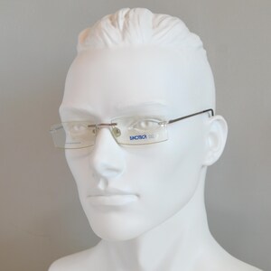 May include: Rimless eyeglasses with a rectangular shape and thin, dark metal arms. The lenses are clear, and the frame features the word "titanium" on one lens and the brand name "Luxotica" on the other. The glasses are displayed on a white mannequin head.