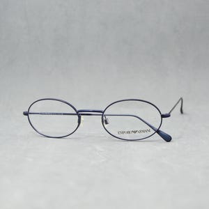 May include: Oval-framed spectacles in dark blue with clear lenses. The brand name "Emporio Armani" is printed on the right lens. The glasses have a minimalist design and are set against a light grey background.