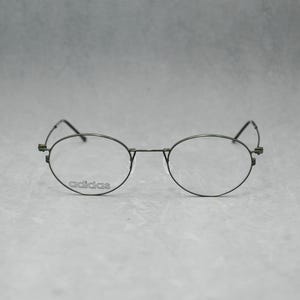 May include: Round-framed eyeglasses with a dark metal finish. The Adidas logo is visible on the left lens. The glasses have a classic, minimalist design, suitable for everyday wear.