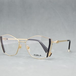 Może przedstawiać: Okulary do czytania w złotej oprawie z czarnymi akcentami i kształtem kociego oka. Soczewki są przezroczyste, a nazwa marki "FURLA" jest nadrukowana na prawej soczewce.