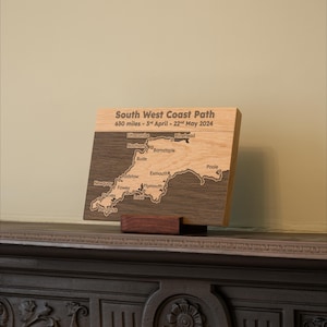 Può includere: Una targa rettangolare in legno con incisa una mappa del South West Coast Path. Il testo recita South West Coast Path, 630 miles - 3rd April - 22nd May 2024, con vari nomi di città. La targa poggia su un piccolo supporto in legno.