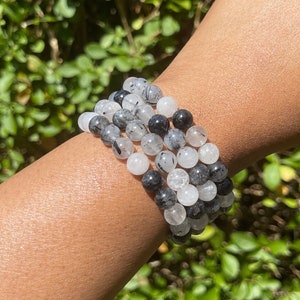 May include: A stack of bracelets made with black, white, and clear round beads. The bracelets are made with natural stones and are worn on a person's wrist.