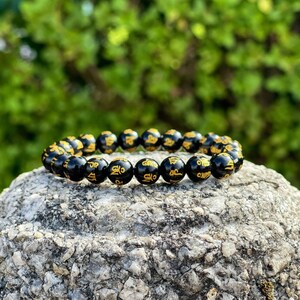 May include: Black beaded bracelet with gold lettering. The beads are round and have a smooth finish. The lettering is in a traditional style.