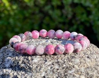 Puede incluir: Un brazalete de piedras preciosas rosa y blanco con motas negras. El brazalete está hecho de cuentas redondas y está apoyado en una roca gris.