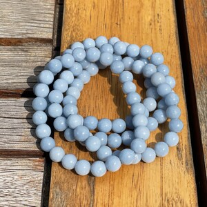 Könnte beinhalten: Ein hellblaues Edelstein-Armband aus runden Perlen. Das Armband besteht aus mehreren Perlensträngen und ist auf einer Holzoberfläche aufgerollt.