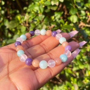 Könnte beinhalten: Ein Armband aus verschiedenen Halbedelsteinen, darunter Amethyst, Rosenquarz und Aquamarin. Das Armband besteht aus runden Perlen und ist auf einem Gummiband aufgefädelt.