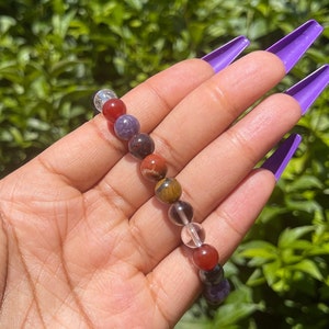 K&ouml;nnte beinhalten: Ein mehrfarbiges Perlenarmband mit klaren, roten, braunen und violetten Perlen. Das Armband besteht aus nat&uuml;rlichen Steinen.