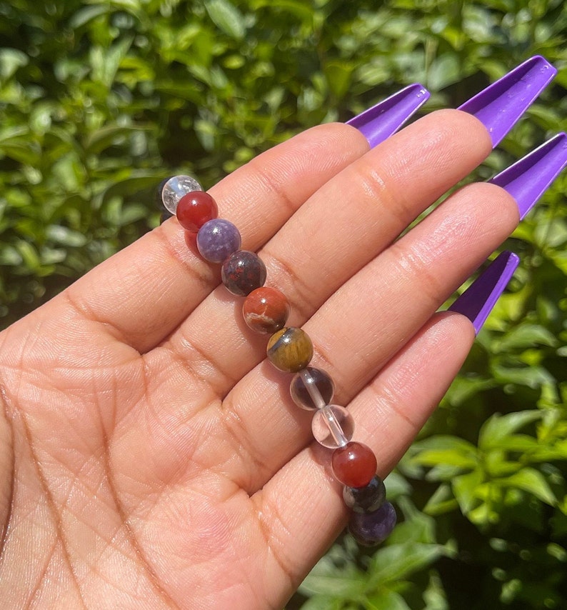 K&ouml;nnte beinhalten: Ein mehrfarbiges Perlenarmband mit klaren, roten, braunen und violetten Perlen. Das Armband besteht aus nat&uuml;rlichen Steinen.