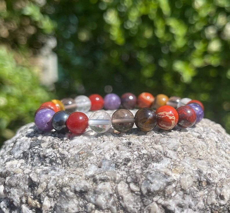 K&ouml;nnte beinhalten: Ein mehrfarbiges Perlenarmband mit roten, orangefarbenen, gelben, gr&uuml;nen, blauen, violetten und schwarzen Perlen. Das Armband besteht aus nat&uuml;rlichen Steinen und liegt auf einem grauen Stein.