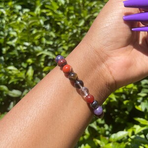 K&ouml;nnte beinhalten: Ein Armband aus bunten Edelsteinperlen. Die Perlen sind rot, orange, braun, klar und lila. Das Armband wird am Handgelenk getragen.