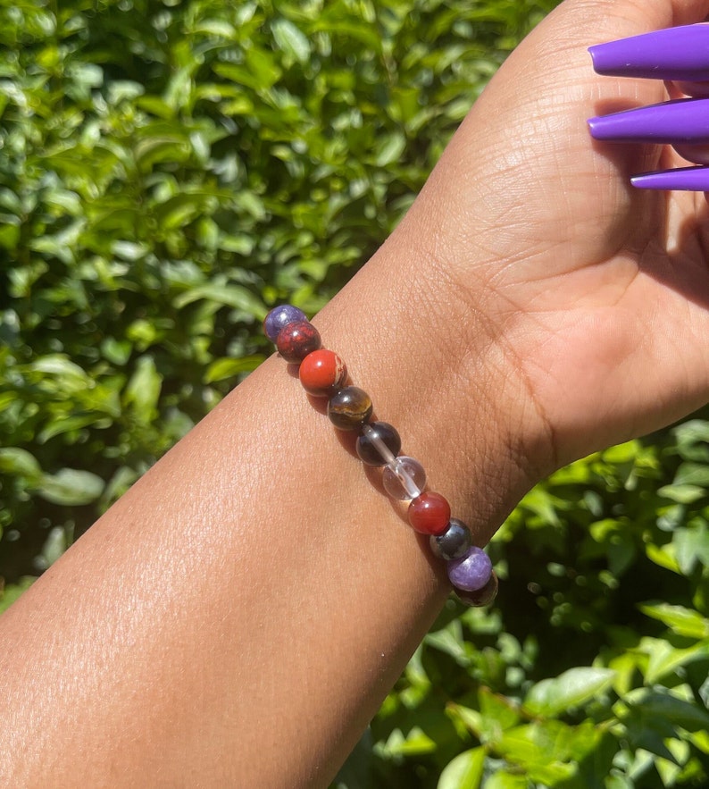K&ouml;nnte beinhalten: Ein Armband aus bunten Edelsteinperlen. Die Perlen sind rot, orange, braun, klar und lila. Das Armband wird am Handgelenk getragen.