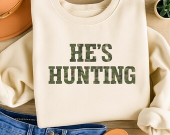 May include: Cream-colored sweatshirt with the text "HE'S HUNTING" in a camouflage pattern. The sweatshirt is folded, with a pair of blue jeans and a brown hat visible in the background. The overall theme is related to outdoor activities.