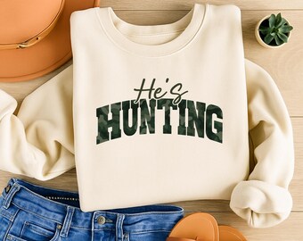 May include: Cream-colored sweatshirt with the text "He's Hunting" in a green camouflage pattern. The sweatshirt is paired with blue jeans and brown sandals. A brown hat and a small potted plant are also visible.