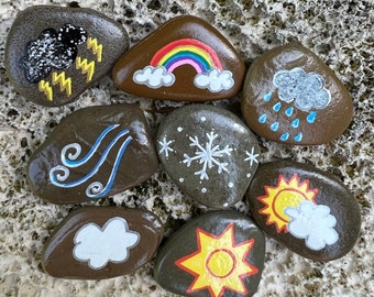Può includere: Nove pietre dipinte che rappresentano diverse condizioni meteorologiche. Le pietre sono dipinte con un arcobaleno, una nuvola con pioggia, una nuvola con fulmini, una nuvola con neve, una nuvola con vento, un sole con una nuvola e un sole con raggi.