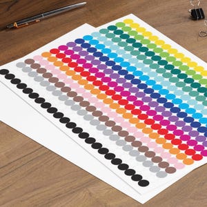 Op de afbeelding: Vellen kleurrijke ronde stickers in een regenboog van tinten, waaronder rood, oranje, geel, groen, blauw en paars. Een zwarte en zilveren pen ligt op het houten oppervlak. De stickers zijn in nette rijen gerangschikt.