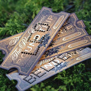 May include: A stack of rectangular paper tickets with a vintage aesthetic, featuring gold and dark brown ornate patterns. The text reads The Polar Express, Round Trip, and Admit One. Each ticket has a series of small punched holes forming a line.