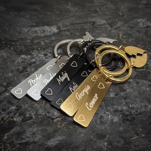 Pode incluir: Uma coleção de chaveiros personalizados em prata, cinza, preto e dourado. Cada etiqueta retangular apresenta um nome e um símbolo de coração. Os chaveiros são presos a argolas de metal, com um pingente em forma de coração.
