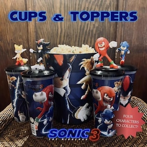 Puede incluir: Colección de vasos y adornos de Sonic the Hedgehog. La imagen muestra varios vasos azules con gráficos de personajes, coronados con figuras. También hay una gran cubeta de palomitas de maíz, junto con el texto "CUPS & TOPPERS" y "SONIC 3 THE HEDGEHOG".