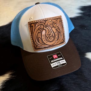 May include: A trucker hat with a white front panel, a brown brim, and a blue mesh back. The hat features a rectangular, embossed leather patch with a detailed floral design. The Richardson 115 Trucker hat has a low-profile fit.