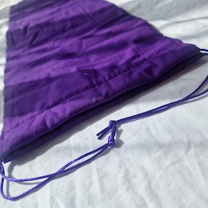 May include: A deep purple drawstring bag, made of a soft fabric, lies on a white surface. The bag has a rounded bottom and a drawstring closure. The fabric has a subtle texture and the drawstring is a matching purple.