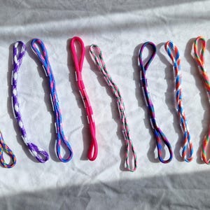 May include: A collection of colourful friendship bracelets, each with a loop at one end. The bracelets display various colour combinations, including pink, purple, blue, and rainbow patterns. They are arranged on a white surface.