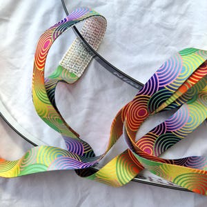 May include: A vibrant headband featuring a repeating circular design in red, orange, yellow, green, and purple. The curved headband is made of fabric. The background is a white sheet.