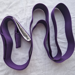 May include: Purple yoga strap with a white lining. The strap is folded and twisted into a figure eight shape.