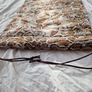 May include: A drawstring bag featuring a snakeskin print in brown, tan, and orange hues. The bag is crafted from a lightweight material and has a dark brown drawstring closure. It rests on a white surface.