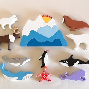 Puede incluir: Un conjunto de figuras de animales de madera, que incluye un oso polar, una morsa, un zorro, un reno, un pingüino, una foca, una ballena, un pulpo y una estrella de mar. Las figuras están pintadas en varios colores, incluyendo blanco, marrón, negro, rojo y azul. El conjunto también incluye una montaña de madera con un océano azul y un sol amarillo.