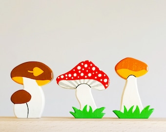 May include: Three wooden mushroom figurines painted in different colors. The first mushroom is brown with a yellow cap and a small brown mushroom on top. The second mushroom is red with white dots and a gray stripe. The third mushroom is yellow with a white stem and green grass at the base.