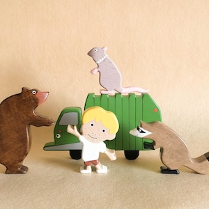 May include: Wooden toy set featuring a brown bear, a raccoon, a gray rat, and a blond-haired child with a green truck. The bear and raccoon are brown, the rat is gray, and the truck is green. The child is wearing a white shirt and brown shorts.