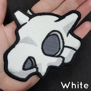 Puede incluir: Parche blanco bordado con forma de calavera, con sombreado gris y contorno negro. El parche tiene la palabra "White" en una fuente manuscrita en la parte inferior. Ideal para chaquetas, bolsos u otros artículos de tela.