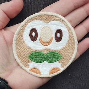Peut inclure: Écusson brodé circulaire représentant un hibou de dessin animé. Le hibou a des yeux bruns, un visage blanc et une forme verte en forme de feuille sous son bec. L'écusson est bordé d'un fil beige et est probablement destiné aux vêtements ou aux accessoires.