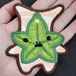 Op de afbeelding: Geborduurde patch met een groen, stervormig blad met een gezicht, tegen een roze en wit gestreepte achtergrond en een bruine rand. De patch heeft zwarte ogen en een zwarte mond.