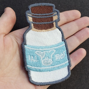 May include: Embroidered patch of a milk bottle. The bottle is white with a blue label featuring a cow graphic and text. The bottle's cap is brown. The patch has a blue border.