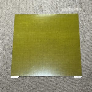 Könnte beinhalten: Eine quadratische, olivgrüne Holzplatte mit strukturierter Oberfläche. Die Platte hat zwei kleine, weiße Laschen an einer Kante. Die Platte ist wahrscheinlich Teil eines größeren Gegenstands, wie z. B. eines Tisches oder eines Regals.