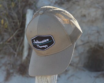 May include: A brown and beige mesh trucker hat with a black and white patch that says "Saltwater Therapy" in white text with a blue line.
