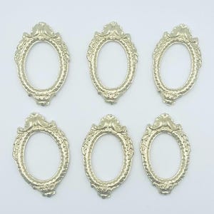 May include: Six ornate, oval-shaped, silver-colored frames. Each frame features a decorative, detailed design around the edges and a crest at the top. The frames are arranged on a white surface.